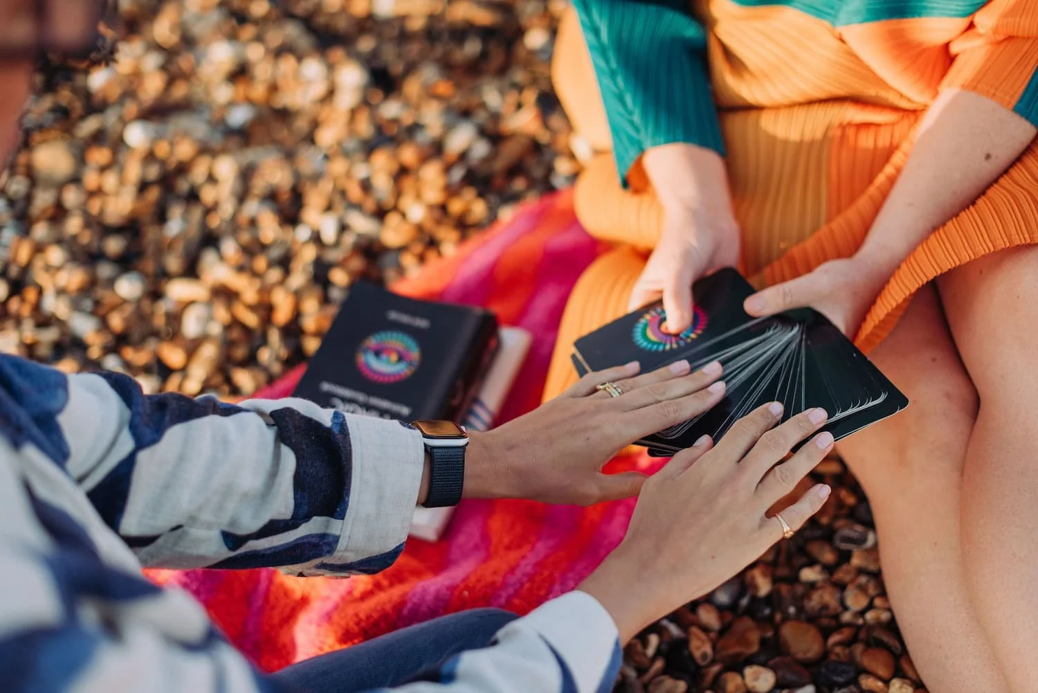 Two people sitting on a colorful blanket on a pebble beach, one of them holding a deck of tarot cards and the other reaching to draw a card, with tarot book nearby.