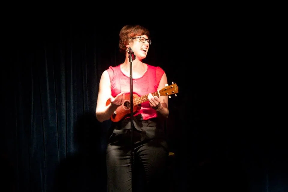 Sarah Weiler with short hair and glasses, wearing a red sleeveless top and black pants, is sitting on a stool on stage, playing a small red ukulele and singing into a microphone, with a dark curtain as the background.