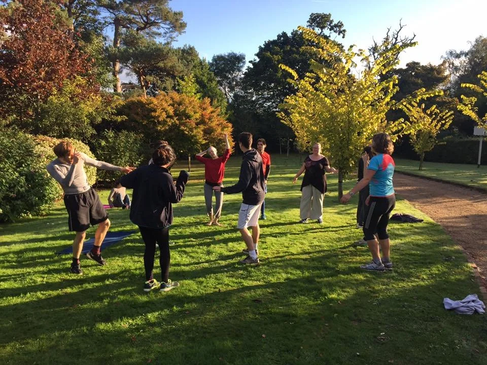 A group of people playing a game of jump rope outdoors on a sunny day, with trees and a clear blue sky in the background.