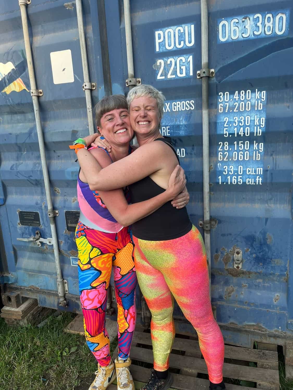 Two women hugging and smiling in front of a blue shipping container at a festival or outdoor event.