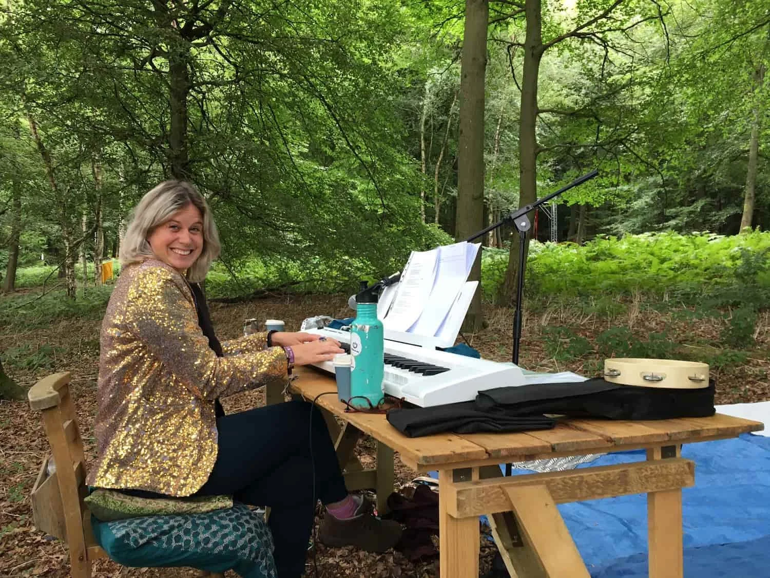 Sarah Weiler with blonde hair wearing a sparkly gold jacket sitting at a wooden table outdoors, playing a keyboard with sheet music, in a forested area.