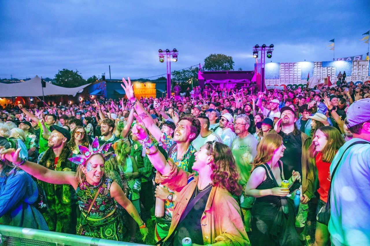 A large crowd of people at an outdoor music festival or concert, dancing and enjoying the event during evening or early night, with stage lighting and sky visible.
