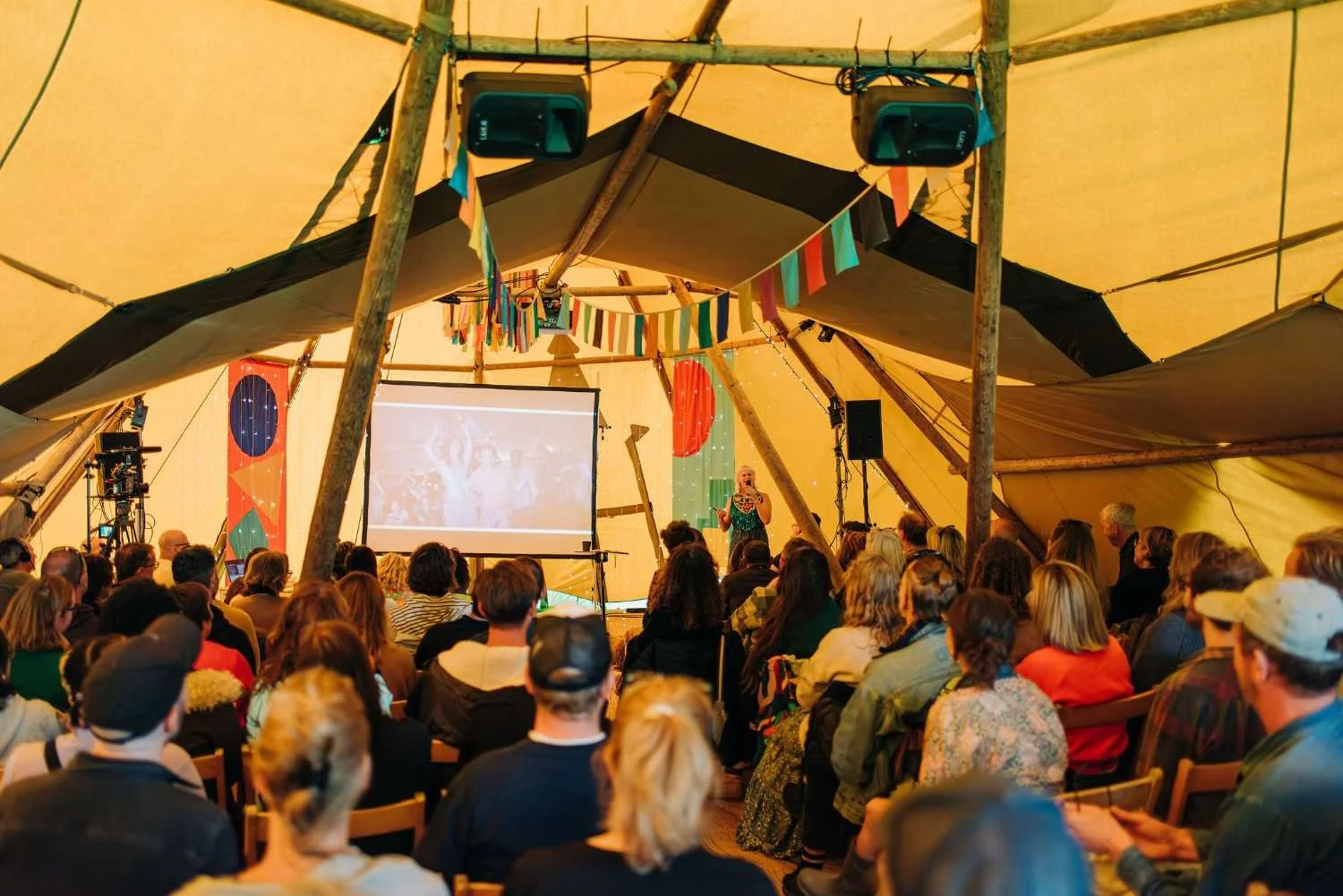 A large group of people seated inside a tent with colorful flags, watching a woman speak on stage with a projector screen behind her.