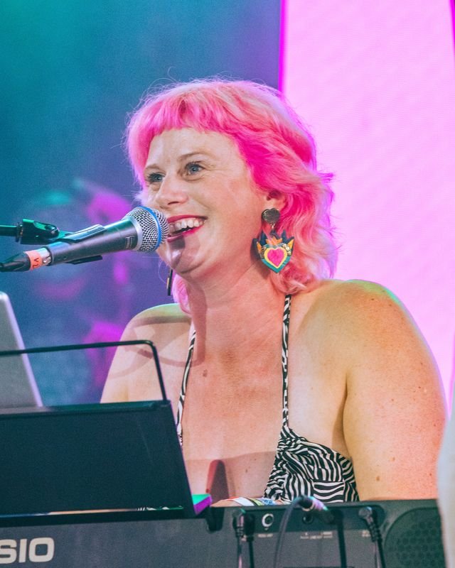 Sarah Weiler with pink hair singing into a microphone while playing a keyboard.