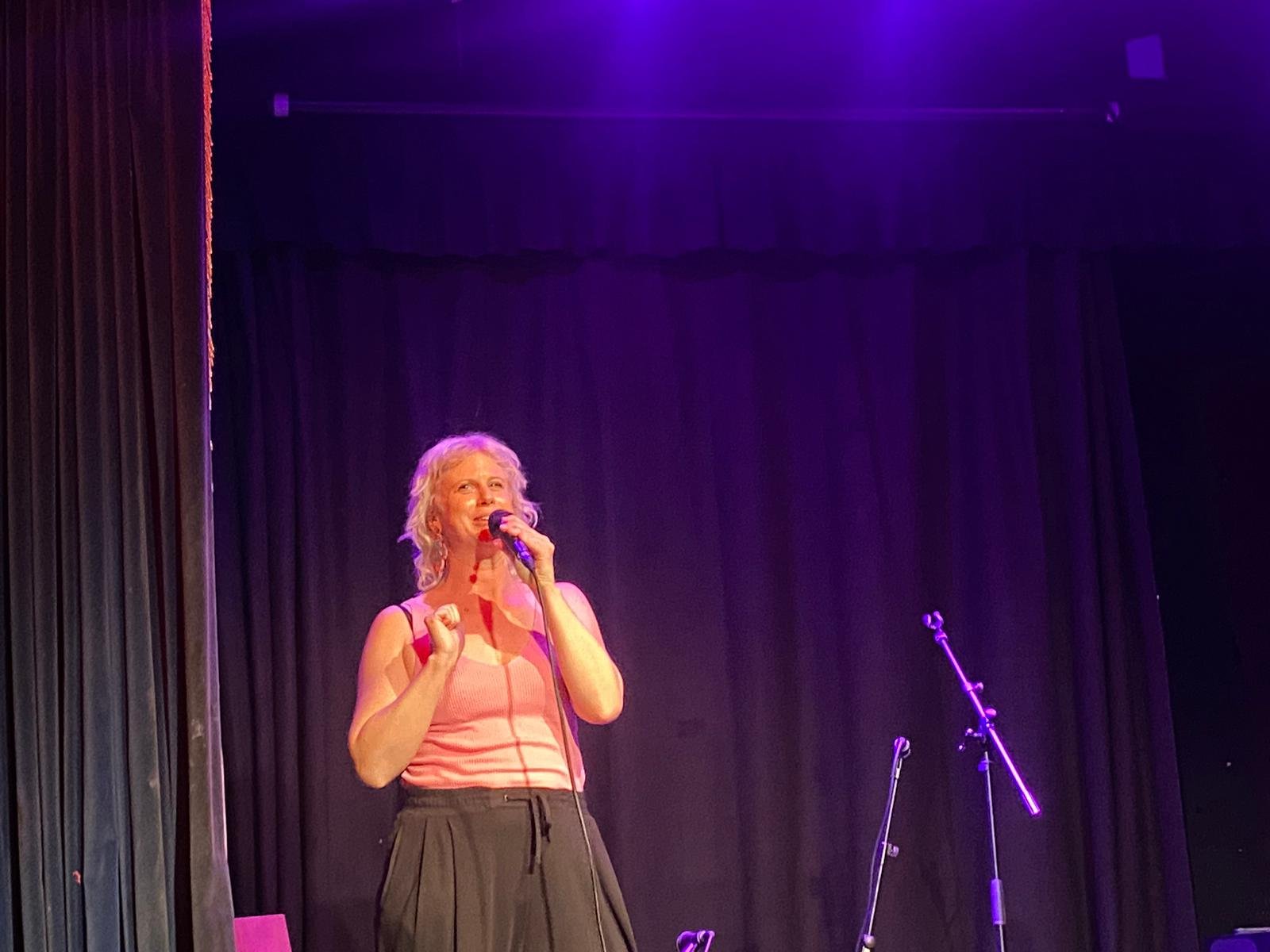 Sarah Weiler with blonde hair singing into a microphone on stage with black curtains and purple lighting.