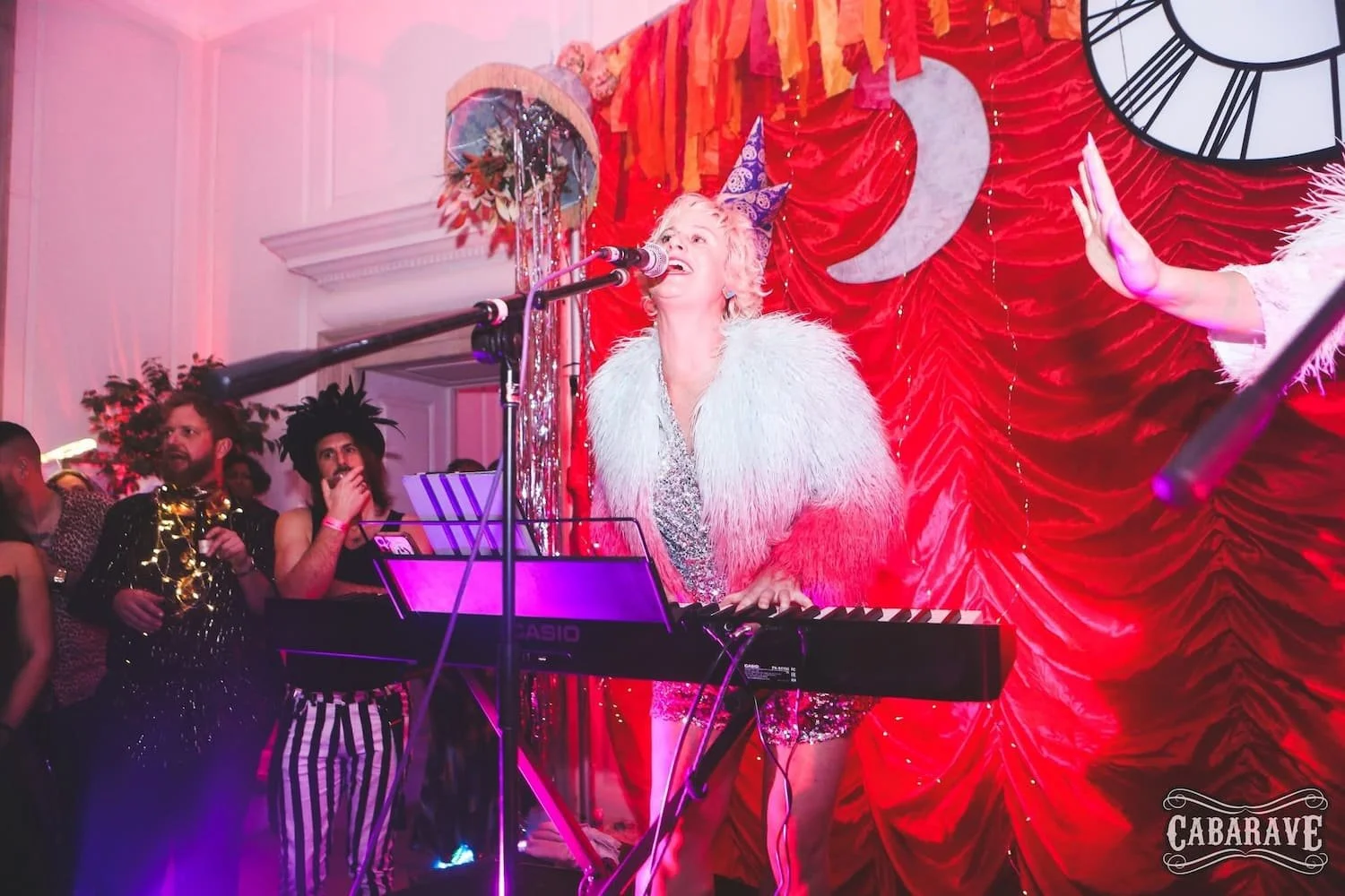 A performer with blonde hair singing and playing a keyboard at a party with a red backdrop and a large clock decoration. The performer is dressed in a shiny outfit with a fluffy white and red coat, wearing a cone-shaped party hat. People in costumes 