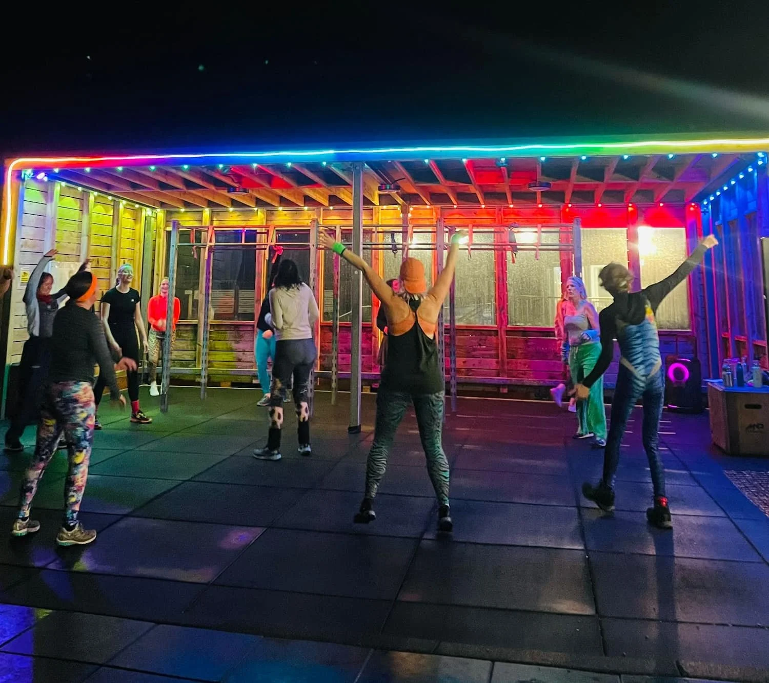 Group of people participating in a dance class or group fitness activity in a colorful, neon-lit outdoor space.