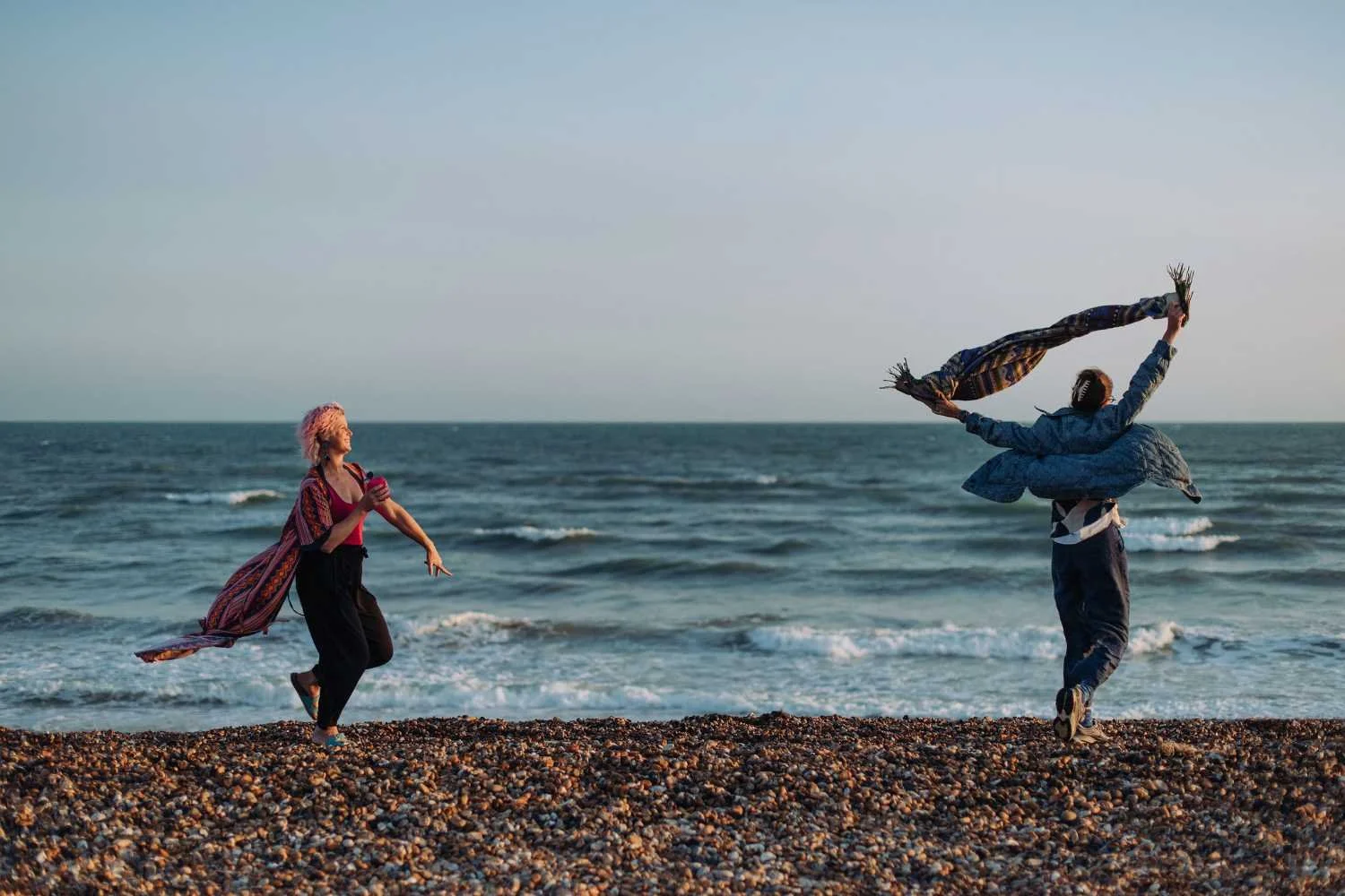 Two people on a pebble beach with the ocean in the background, one holding a phone and the other throwing a scarf into the air, both wearing casual clothing.