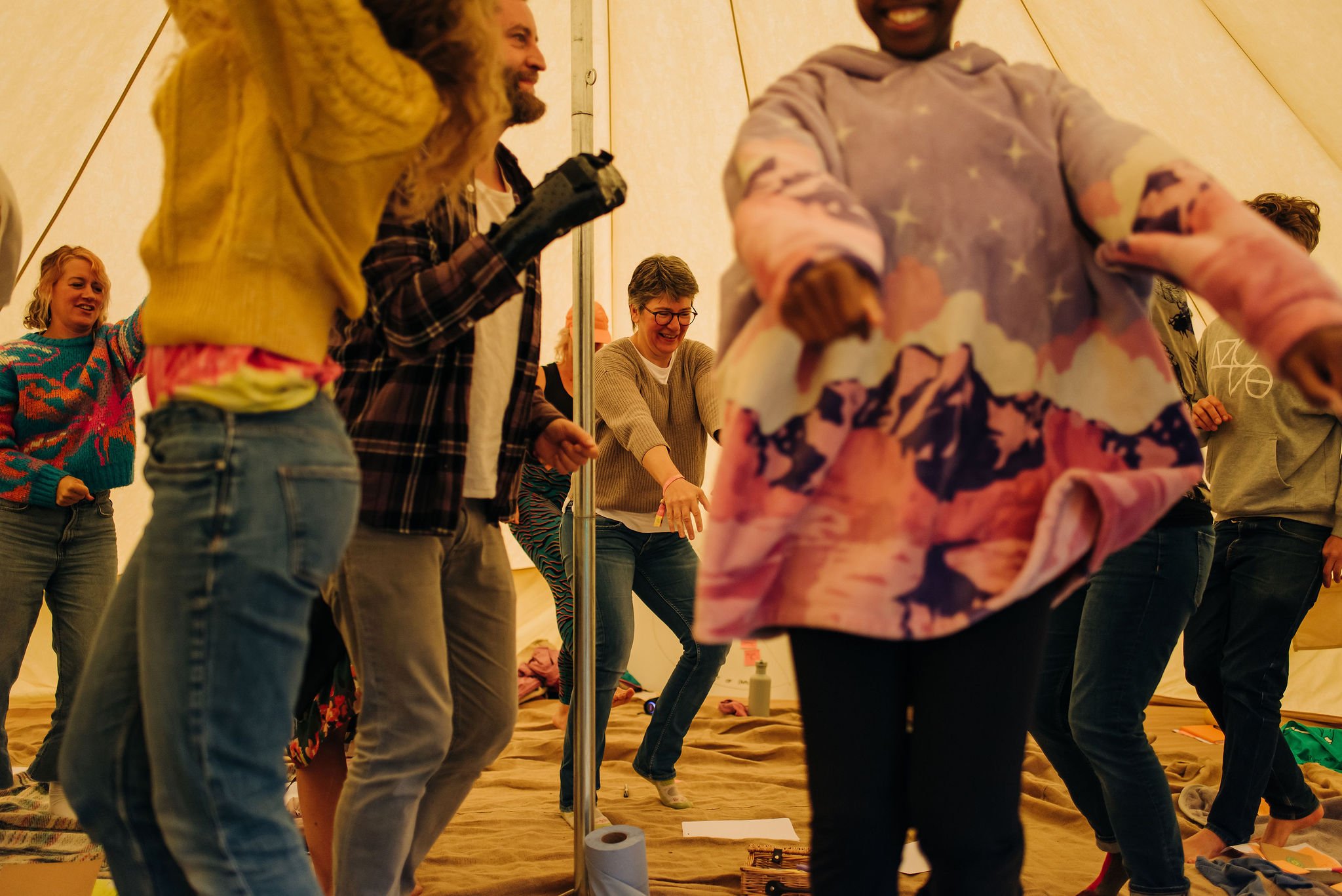 People standing in a circle inside a large tent, holding hands and smiling, with yoga mats and supplies on the floor.