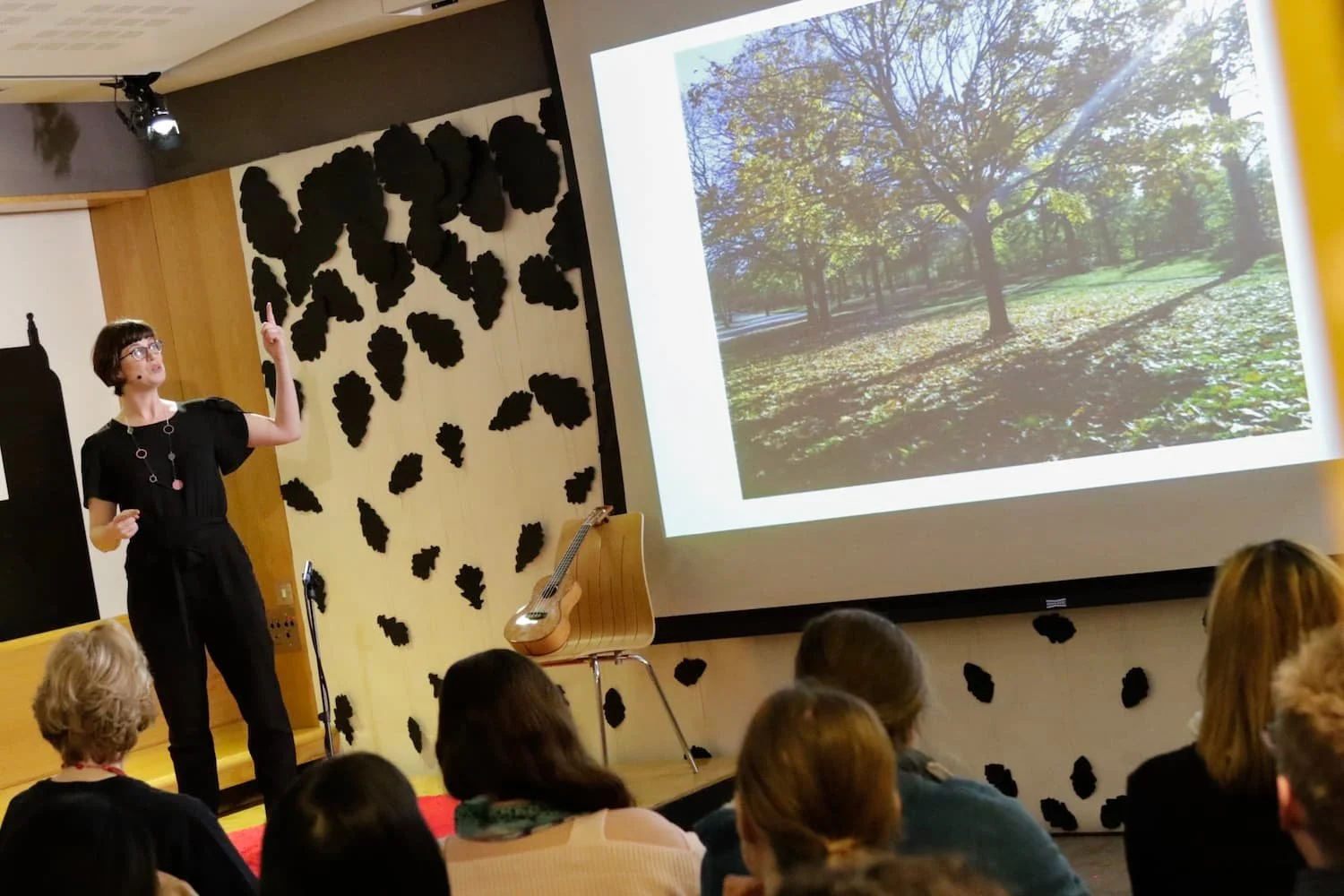 Sarah Weiler giving a presentation in a conference room with an audience. She is pointing to a large screen showing a photo of trees in a park. The room has decorations on the wall resembling black leaves, and a guitar is placed on a chair.