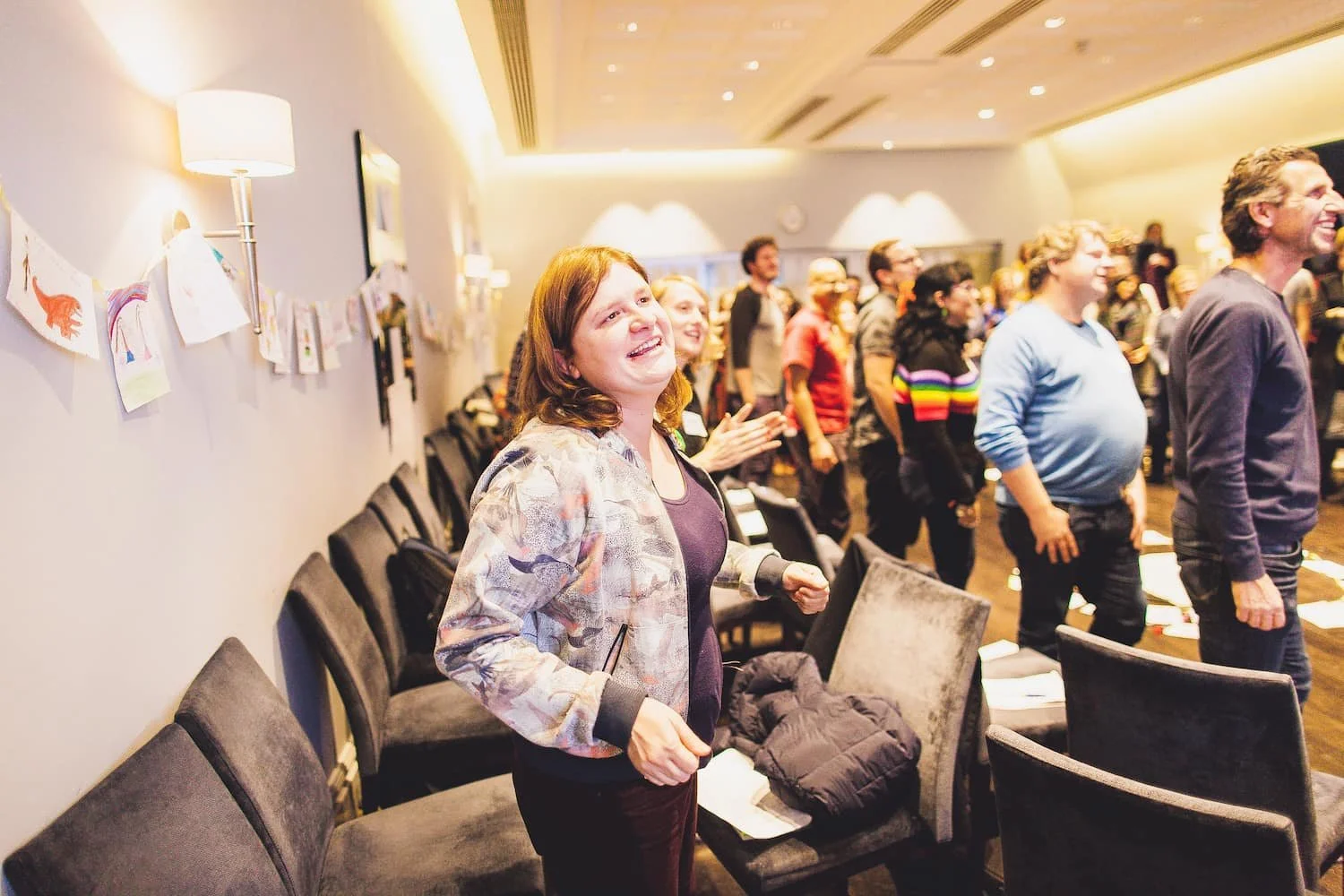 People standing and smiling at a gathering or event in a room with walls decorated with children's artwork and seating chairs.