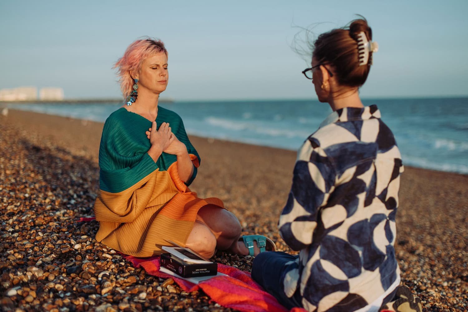 Two women on a pebble beach during sunset, one sitting cross-legged with hands on her chest, eyes closed, wearing a colorful dress and large earrings; the other kneeling with a clip in her hair, wearing glasses and a patterned shirt, facing her.