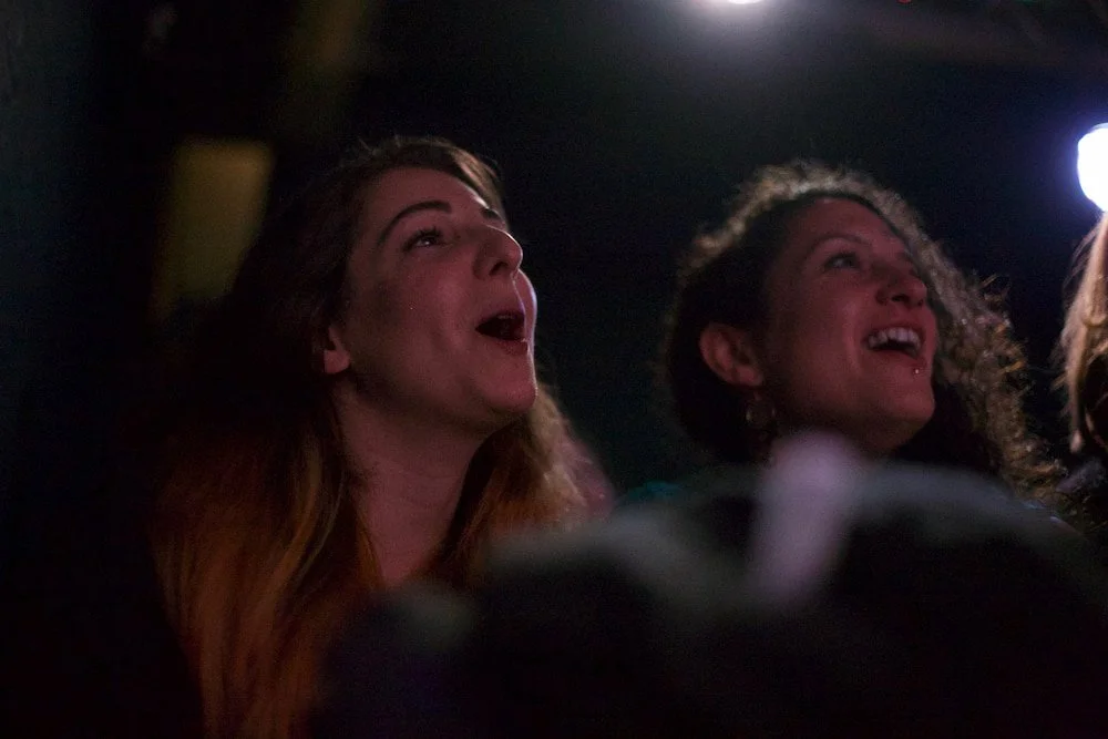 Two women at an event, appearing emotional and joyful, with one singing or shouting and both smiling.