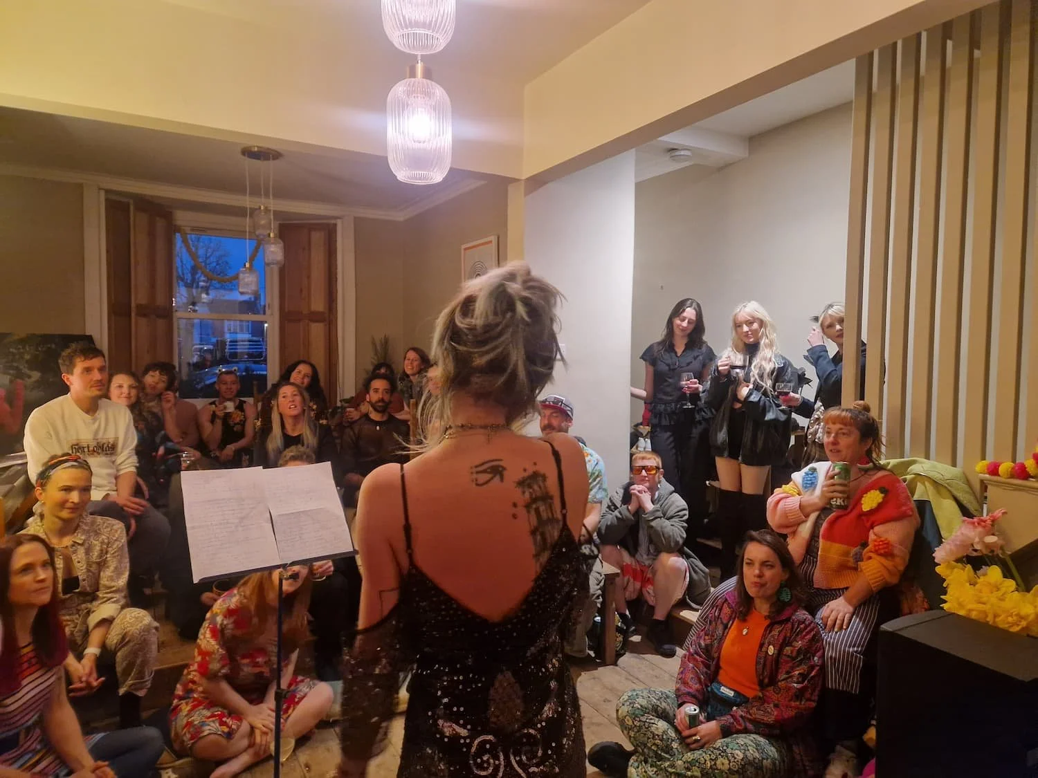 A woman with tattoos and a dress is performing or speaking to a group of people at an indoor gathering or party. The audience is seated and standing, enjoying the event. The room has warm lighting and a window in the background.
