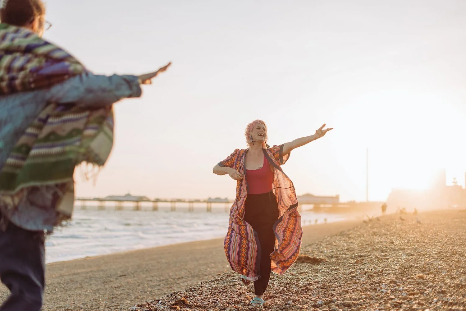 Sarah Weiler with pink hair wearing a colorful dress and red top, smiling and reaching out her arm on a beach at sunset while a person in a patterned jacket gestures towards her from the left.