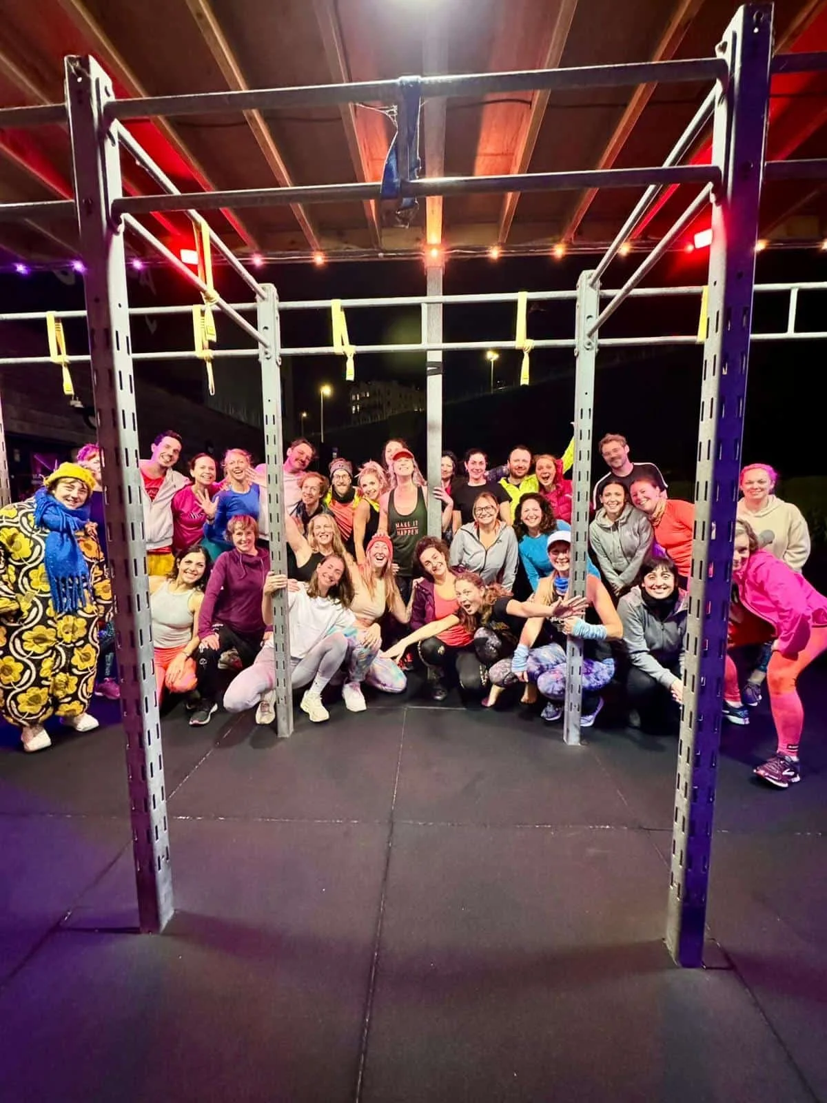 Group of people in workout clothes posing together behind a metal fitness rack in a gym or fitness studio at night.