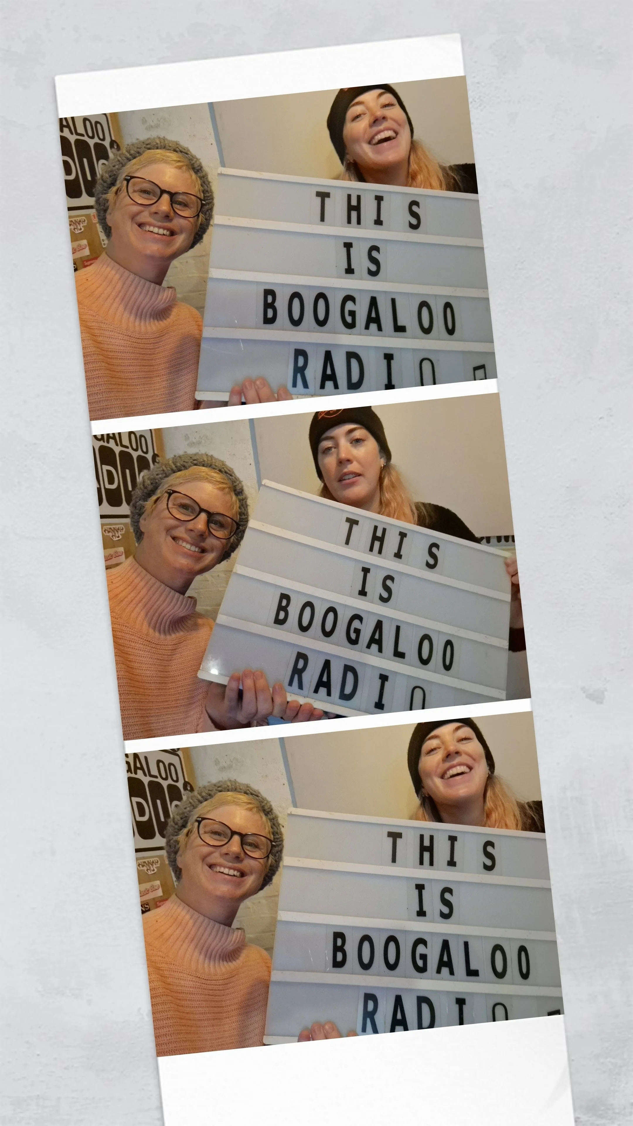 Two women smiling and holding a lit-up sign with the message 'This is Boogallo Radio.' The woman on the left wears glasses and a pink sweater, while the woman on the right wears a black beanie and black shirt.