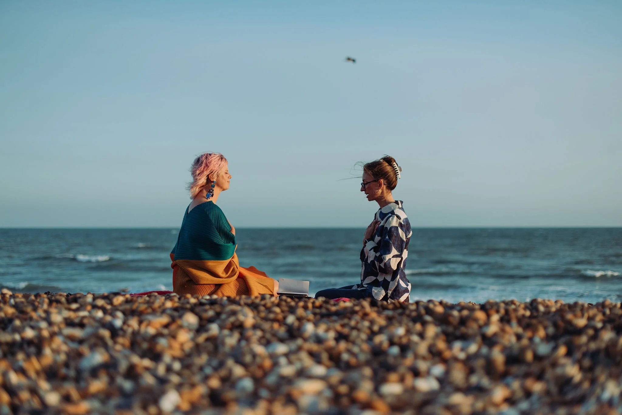 Two women sit on a rocky beach facing each other with the ocean in the background under a clear blue sky. One woman has pink hair and wears a blue top with orange pants, while the other has brown hair tied back with a hair clip and wears glasses and 