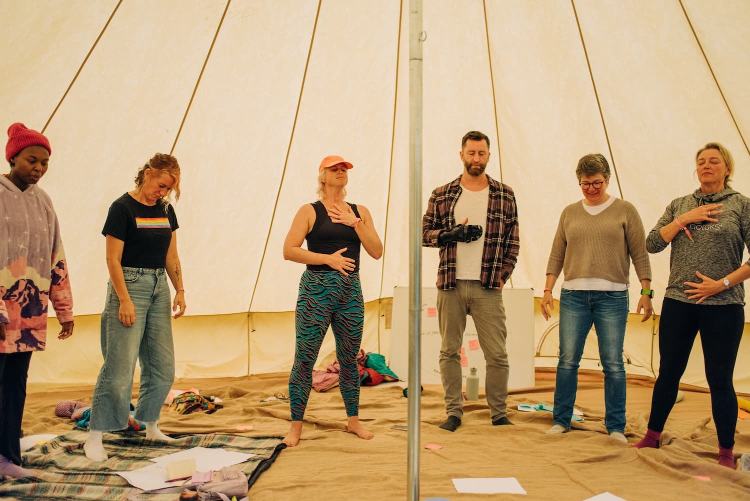 Six people stand inside a large beige tent, appearing to engage in a spiritual or meditative activity. They have their eyes closed, some with their hands on their chest or stomach, and expression of concentration or calm. The tent’s interior is simple, with scattered papers and personal items on the ground.