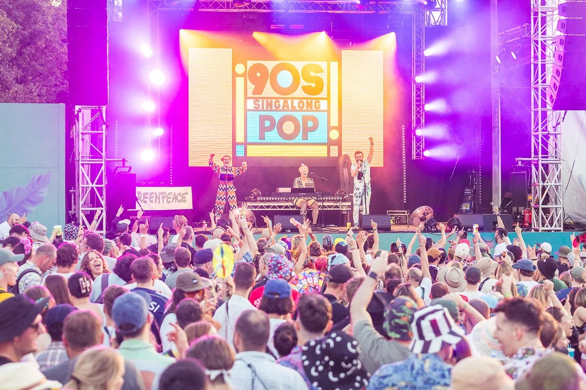 Crowd watching a live outdoor concert at the 2019 Sing Along Pop event, with a stage featuring performers and a large screen displaying the event logo, illuminated by colorful stage lights.