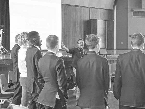 A group of six people, mostly men in suits, standing and facing a woman who appears to be speaking or giving instructions in a room with a stage and chairs.