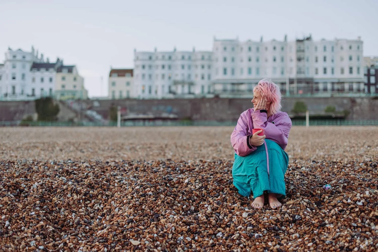 Sarah Weiler with pink hair sitting on a pebble beach, holding a pink cup and looking to the side with a smile. She is dressed in a colorful jacket and pants, with clouds and white buildings in the background.