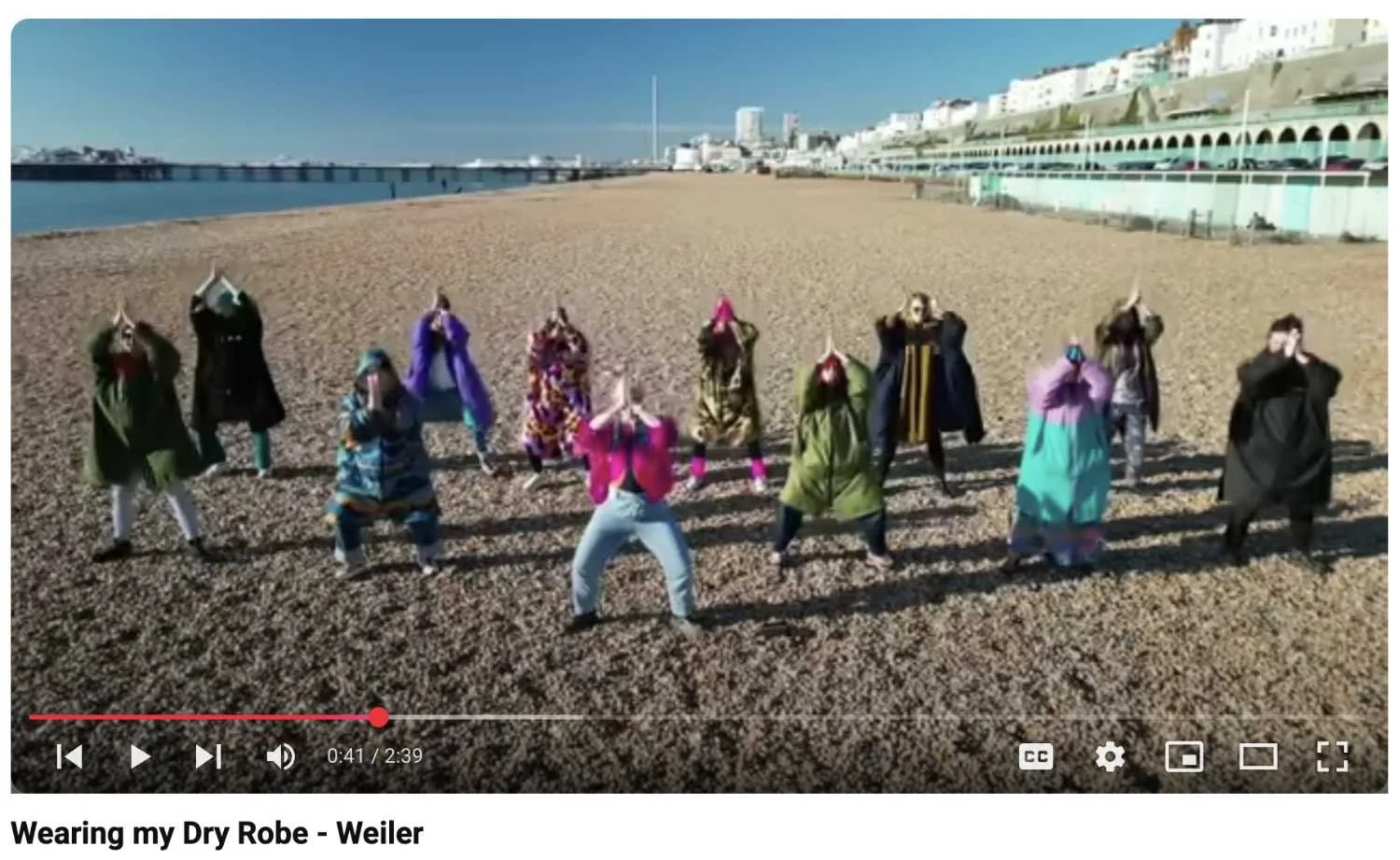 Group of people on a beach wearing various colorful outfits, standing on sand with buildings and a pier in the background.