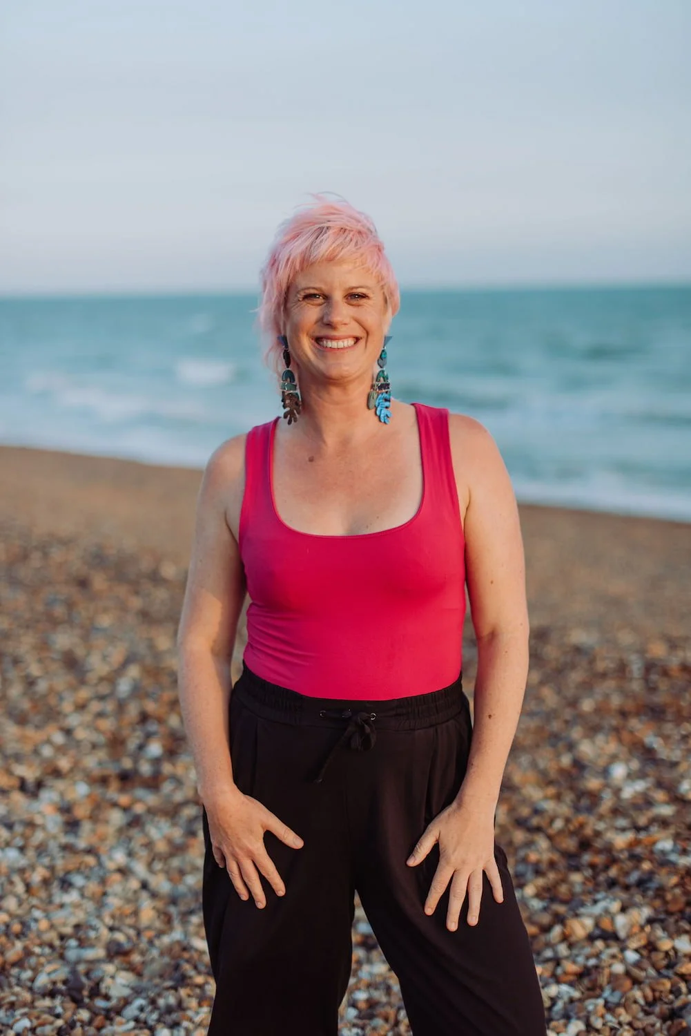 Sarah Weiler with short pink hair smiling at the camera on a pebble beach with the ocean in the background.