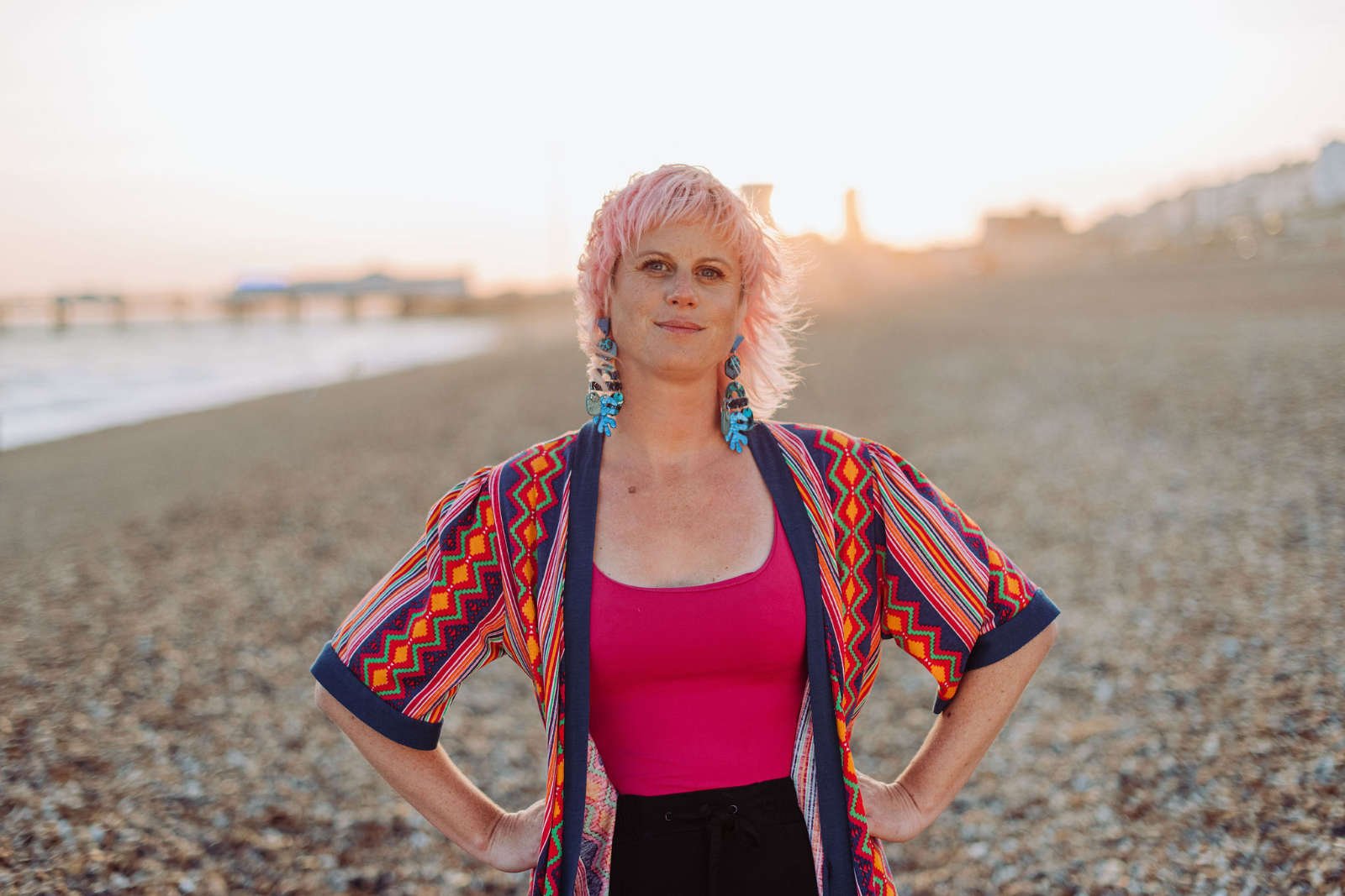 Sarah Weiler with pink hair and colorful earrings standing on a beach during sunset, wearing a vibrant, patterned kimono over a pink top.