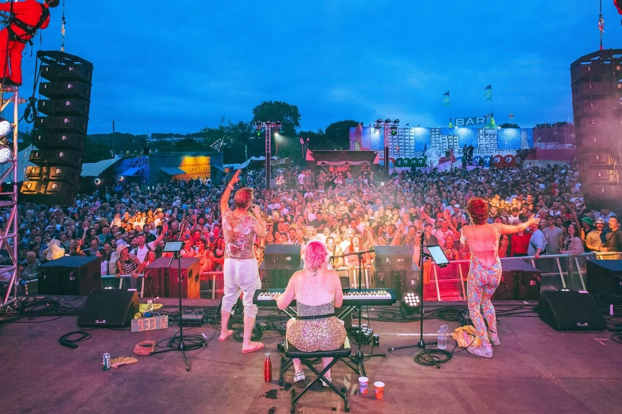 A live music performance on stage with three musicians and a keyboardist facing a large, standing crowd during dusk. The stage has various sound equipment and colorful lighting, creating a vibrant festival atmosphere.