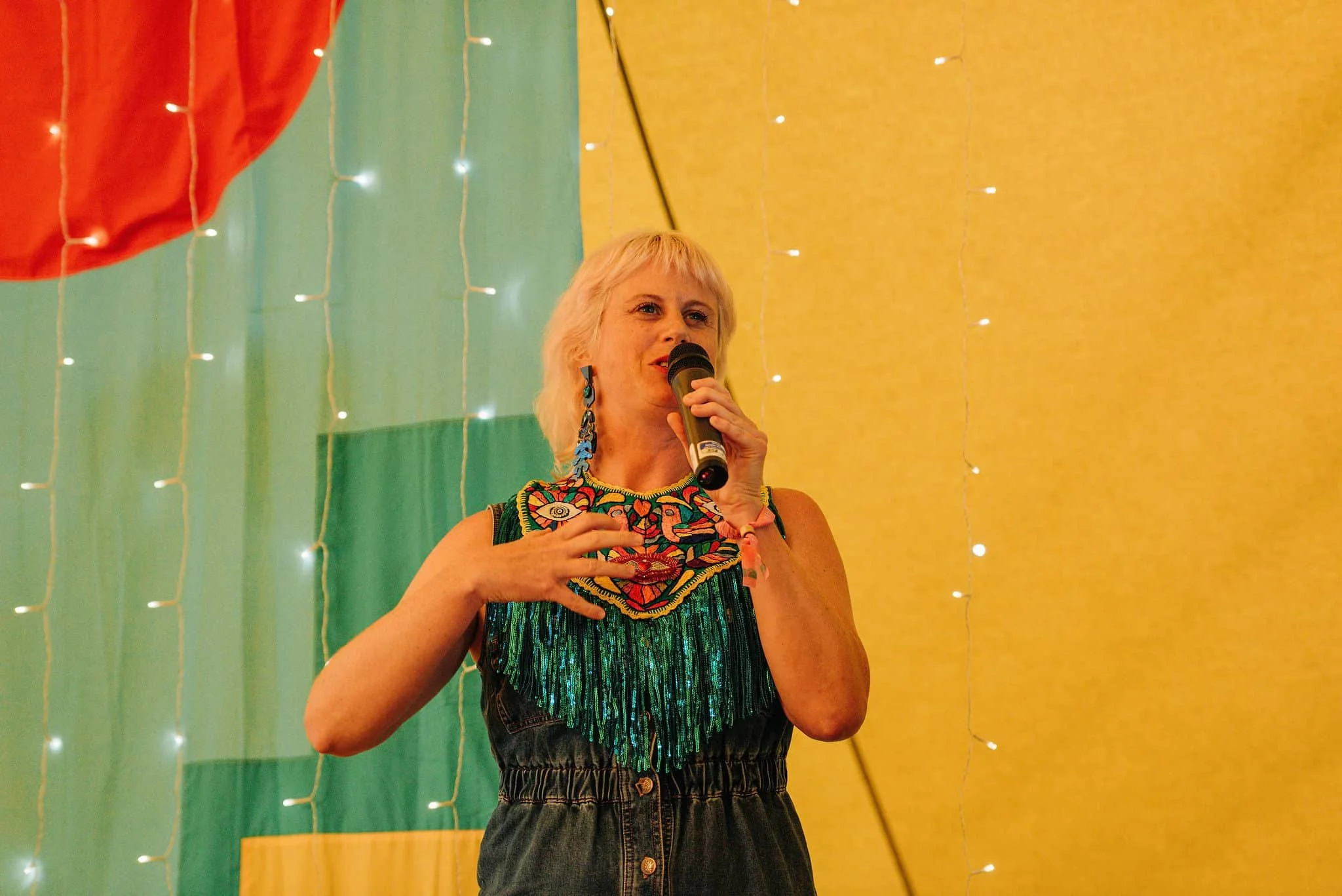 Sarah Weiler with short blond hair wearing colorful earrings and a multicolored embroidered top, speaking into a microphone against a yellow and green backdrop decorated with fairy lights.