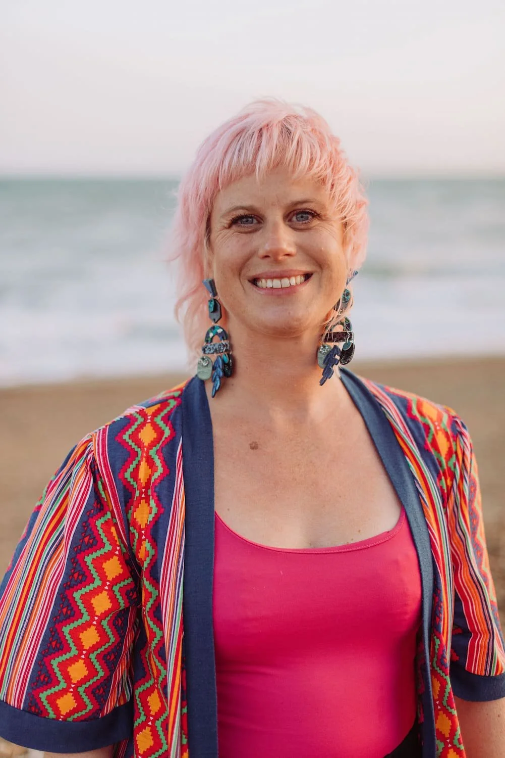Sarah Weiler with pink hair smiling at the camera on a beach, wearing colorful earrings, a pink top, and a vibrant, patterned jacket.