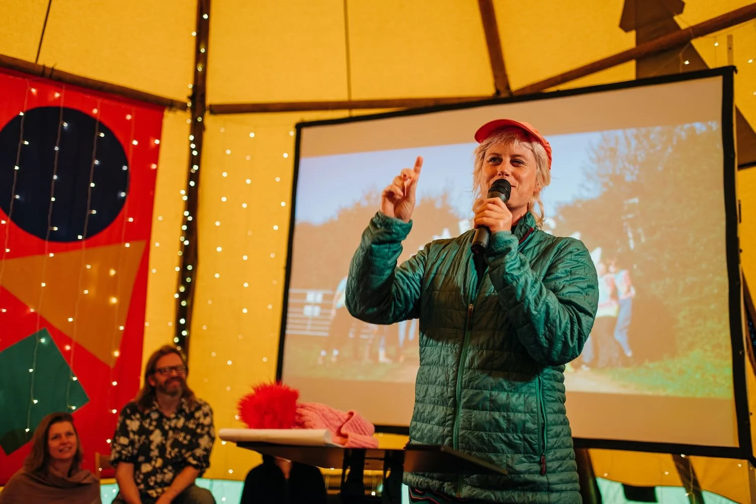 Woman with short gray hair wearing a red hat and green jacket speaking into a microphone at an indoor event with a large screen and a group of people watching.
