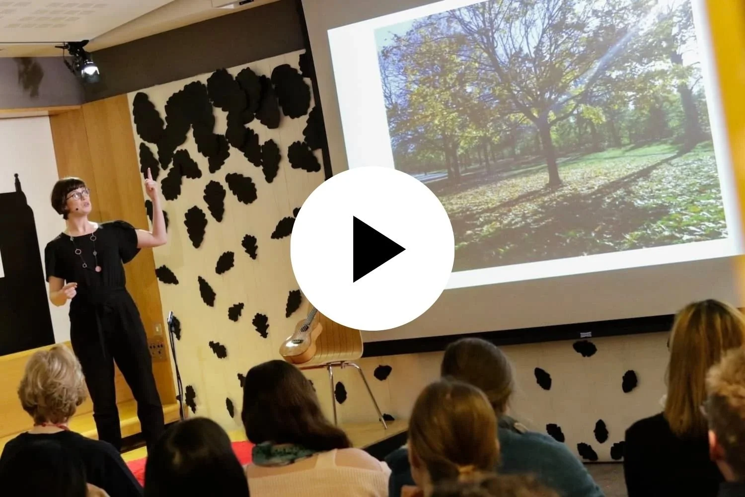 Sarah Weiler giving a presentation to an audience in a conference room. The slide shows a landscape with trees and sunlight.