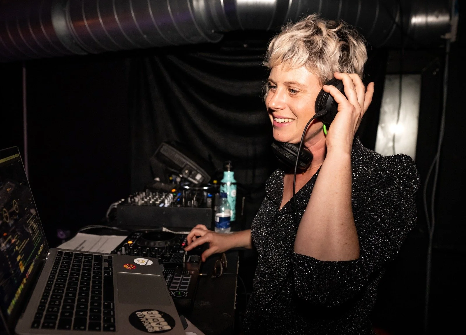 Sarah Weiler with short blonde hair smiling as she wears headphones while working at DJ equipment and a laptop in a dark room.