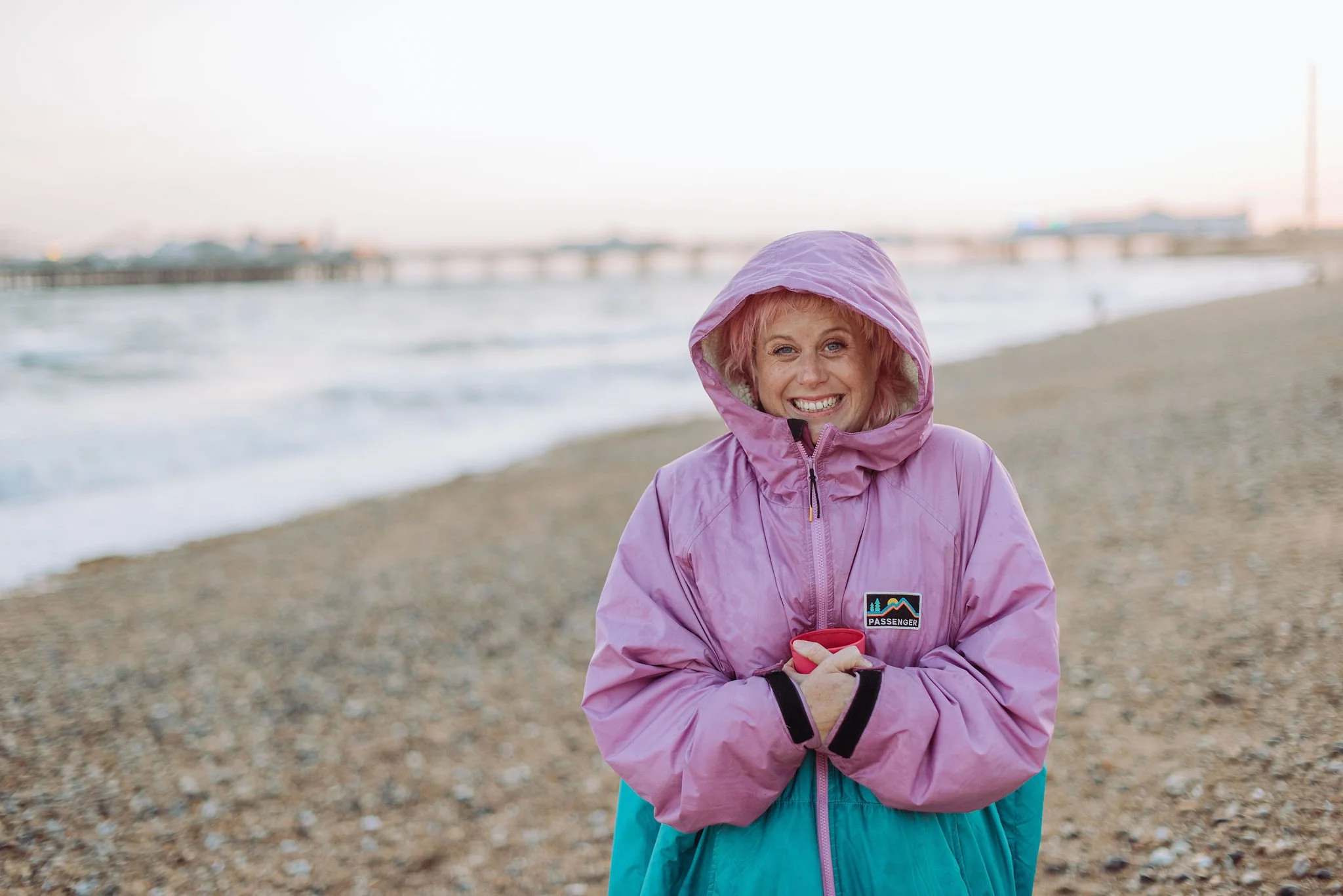 Sarah Weiler smiling in a pink waterproof jacket with a hood, standing on a beach with a pier and water in the background.