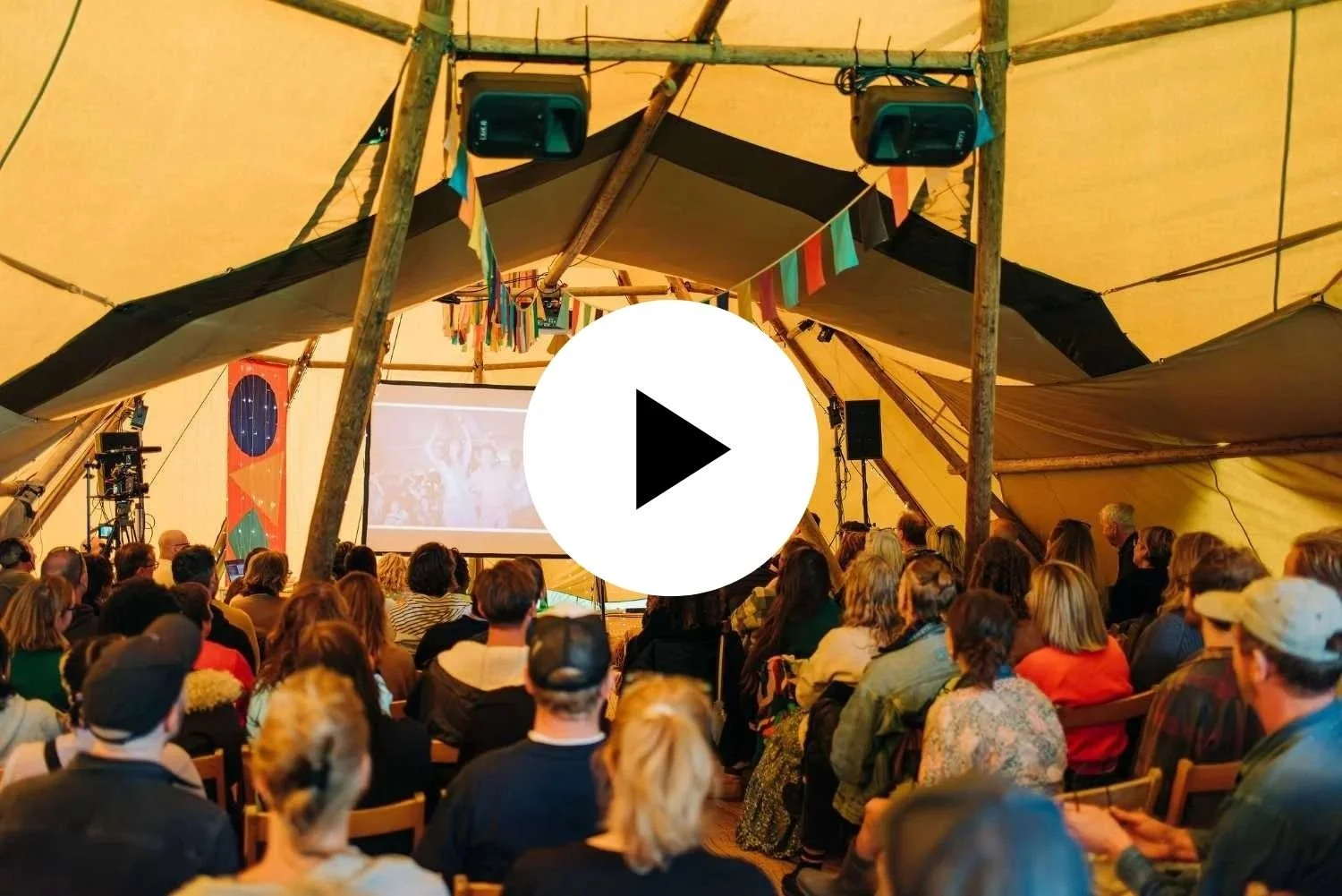 Crowd of people sitting under a large tent watching a presentation projected on a screen