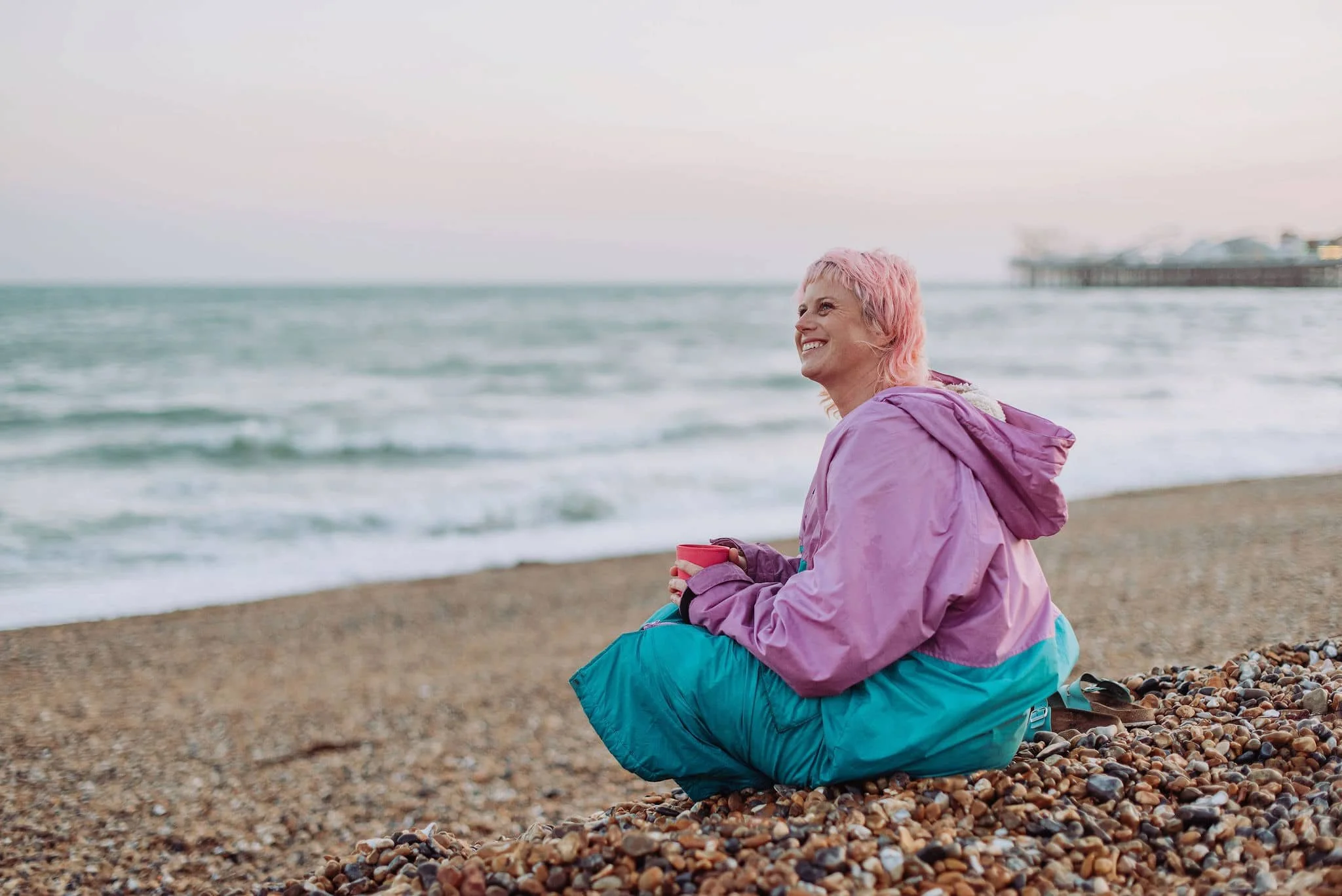 Sarah Weiler with pink hair wearing a purple and teal jacket sitting on a pebble beach, holding a red cup, smiling while looking out at the sea with a pier in the background.