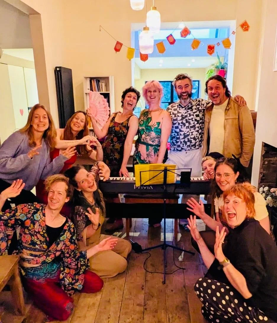 Group of twelve people gathered around a keyboard in a warmly decorated room, celebrating with smiles and gestures, with colorful bunting hanging from the ceiling.