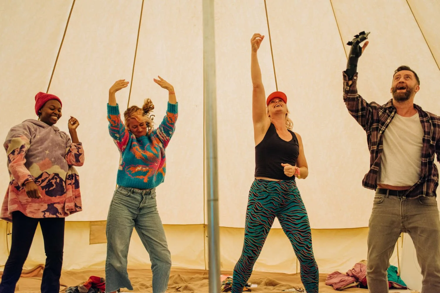 Four people dancing and singing inside a large tent with beige fabric walls. The group includes two women and one man, all with joyful expressions, dressed casually in colorful and patterned clothing.