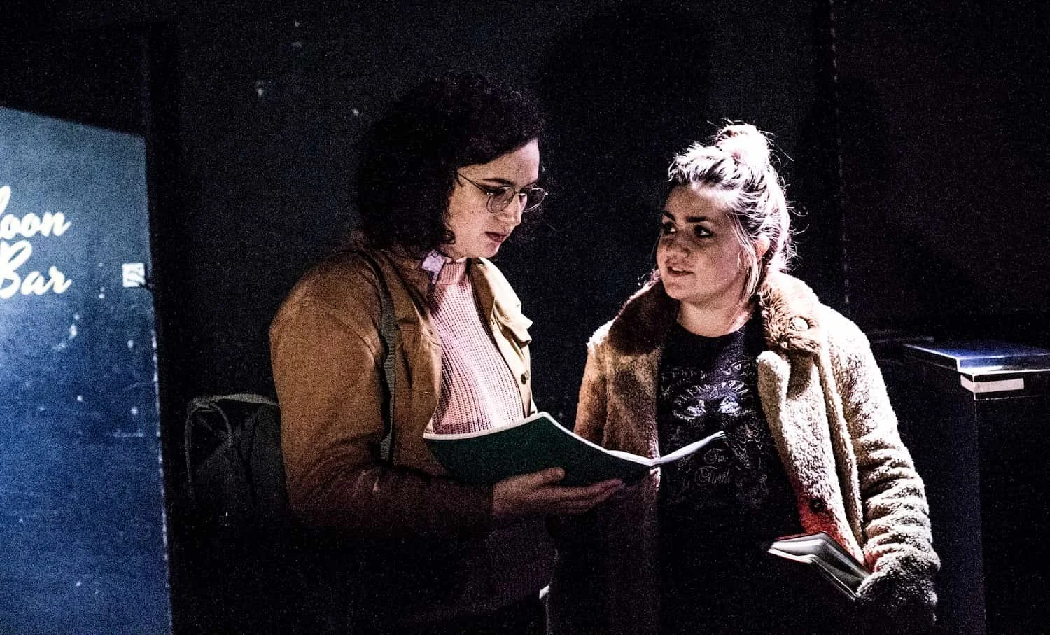 Two women standing and looking at a notebook in a dark room, with a sign in the background that reads 'Room Bar'.