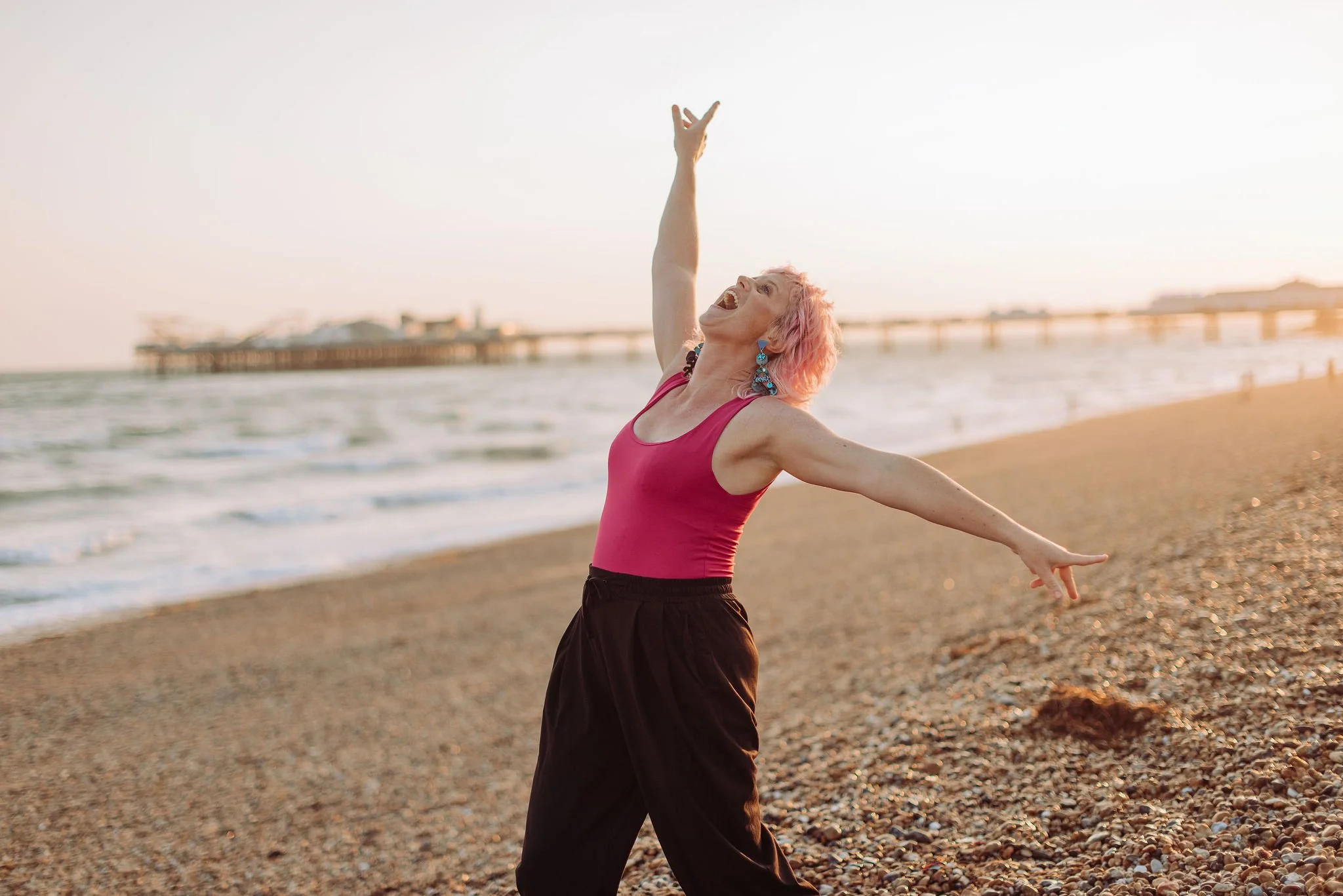 Sarah Weiler with pink hair dancing joyfully on the beach at sunset, with arms outstretched and head tilted back.
