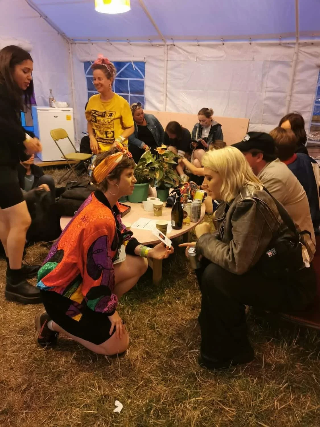 Group of people gathered inside a white tent, sitting and standing around a table with drinks and snacks, engaging in conversation.