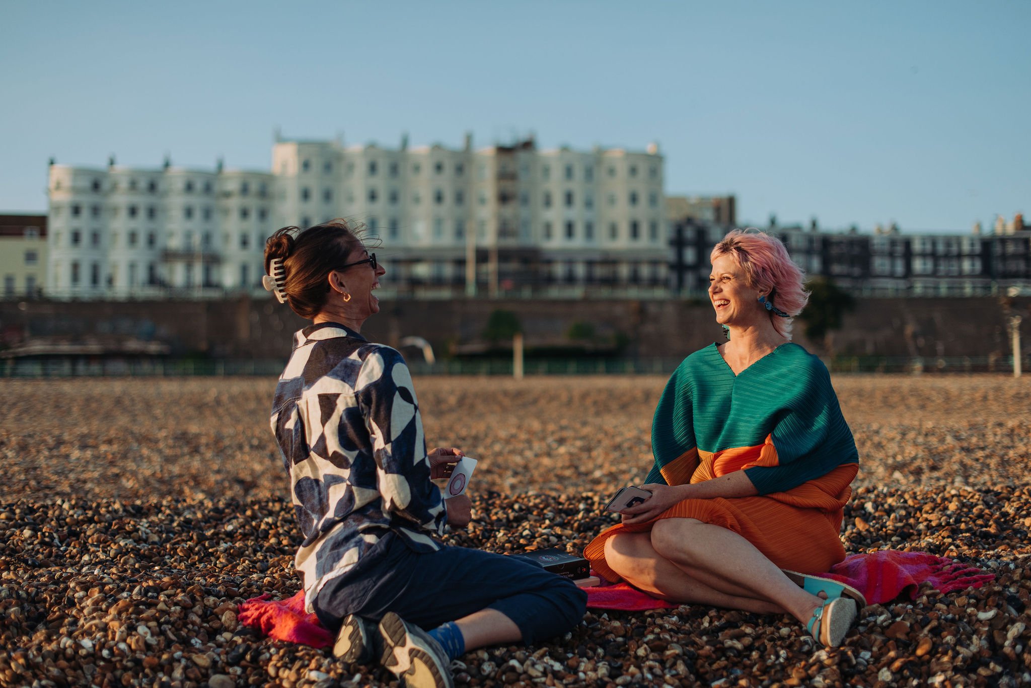 Two women sitting on a pebble beach, laughing and talking, with a cityscape of white and dark apartment buildings in the background during daylight.
