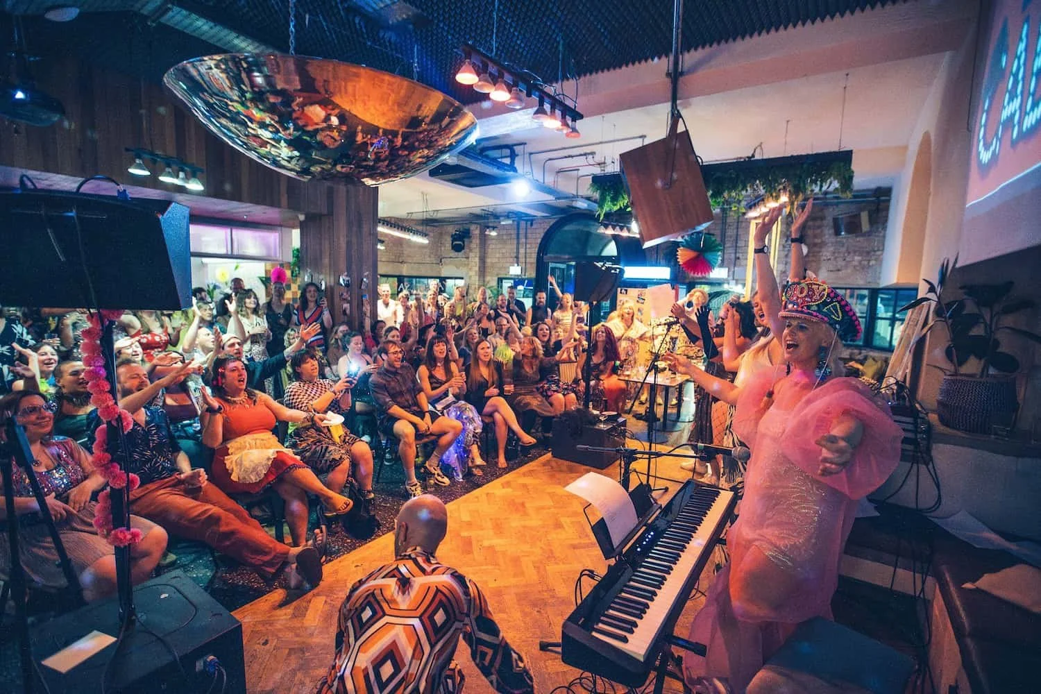 A lively indoor celebration with a diverse crowd watching performers on stage. The performers, dressed in colorful costumes, are singing and dancing with one raised arm. The audience appears engaged and excited, with some taking photos. The setting has warm lighting, a wooden floor, and a ceiling decorated with hanging objects and greenery.