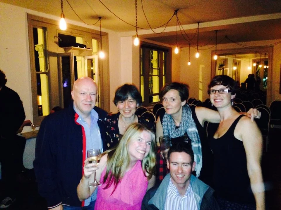 Group of six people celebrating, inside a warmly lit room with hanging light bulbs and windows.