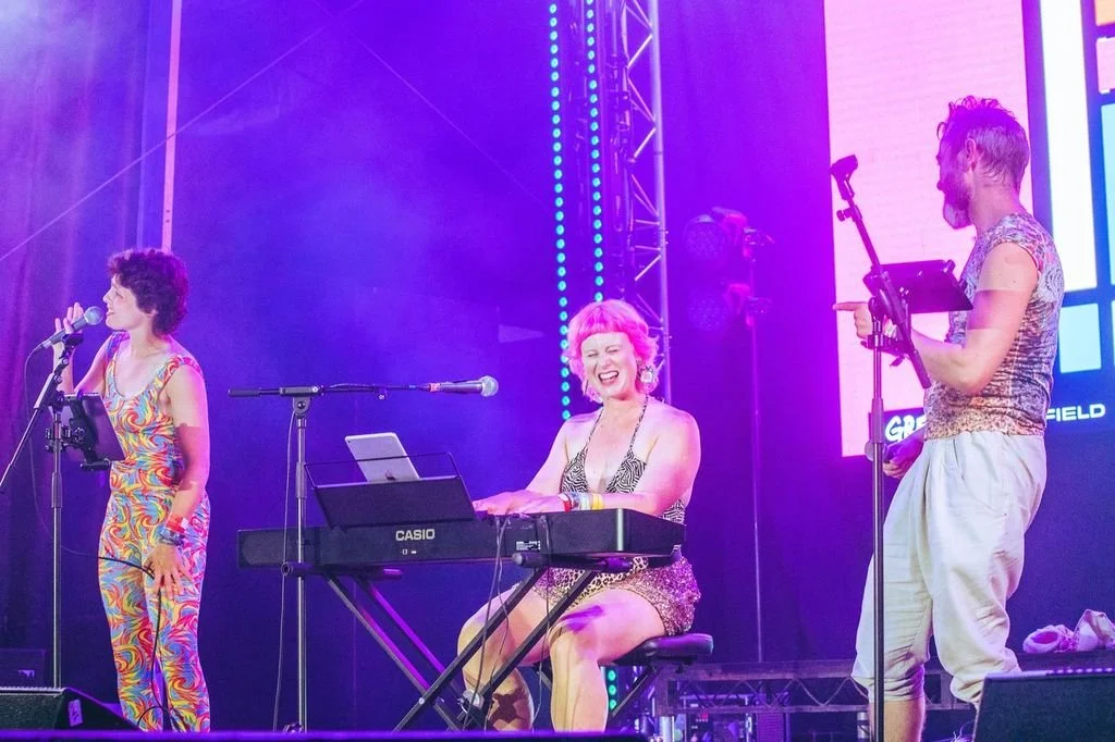 A musical performance on stage with three performers, one playing keyboard, one singing into a microphone, and one reading from a tablet, with colorful stage lighting.
