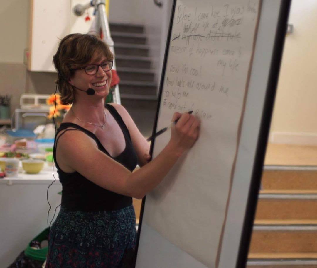 Sarah Weiler with short brown hair, glasses, and a headset smiling as she writes on a whiteboard in a classroom or workshop setting.