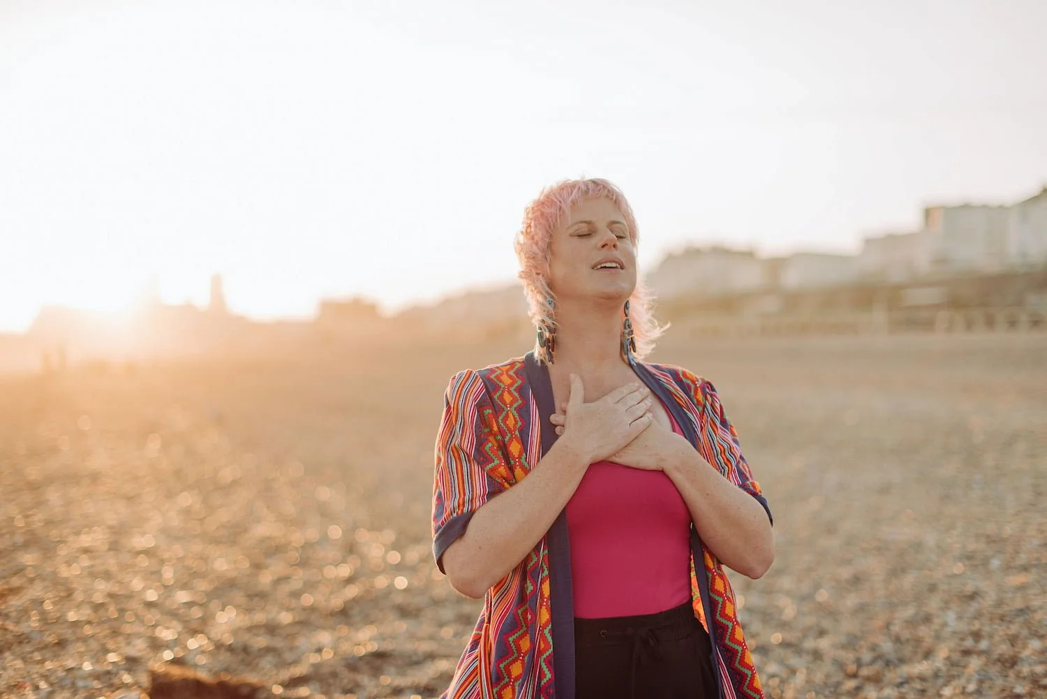 Sarah Weiler with pink hair and colorful clothing standing on a beach at sunset, eyes closed, holding her hand to her chest.