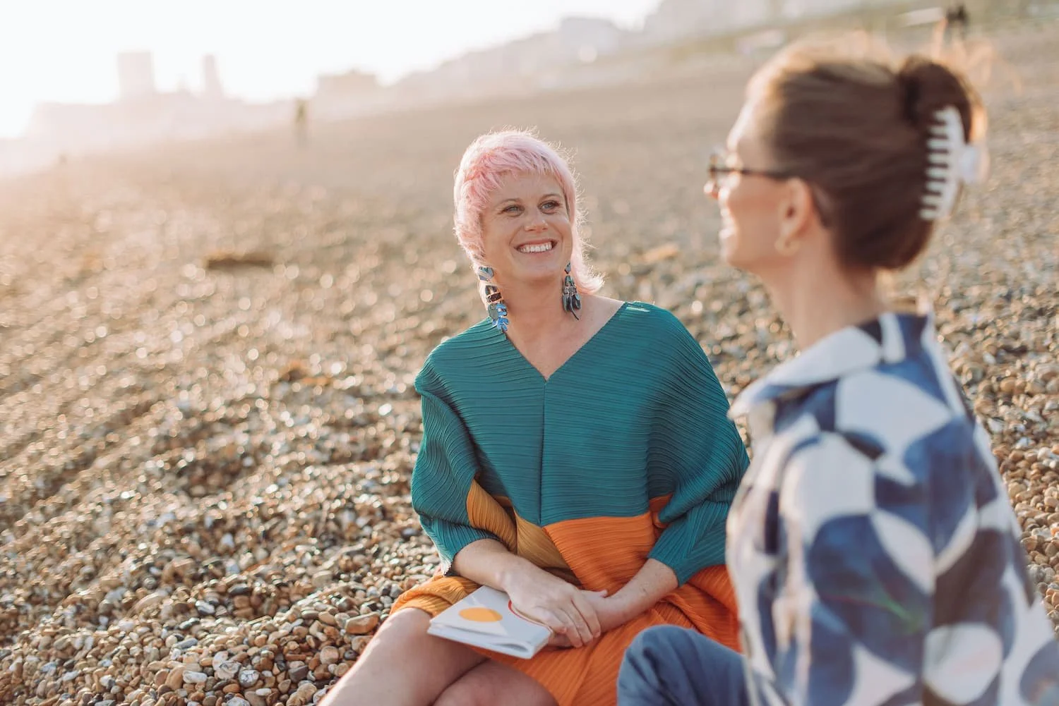 Two women sitting on a pebbled beach, smiling and talking. One woman has pink hair, wearing a teal and orange dress, holding a notebook. The other woman has brown hair styled in an updo with a hair clip, wearing glasses and a patterned shirt.